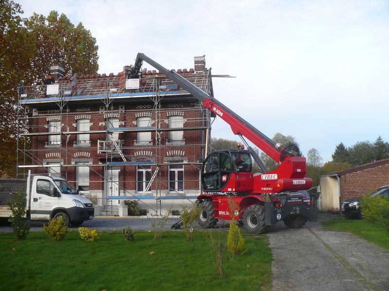 Un camion-grue rouge soulève des matériaux pour monter sur le toit d'une maison en briques en cours de réparation, une camionnette blanche se trouvant à proximité.