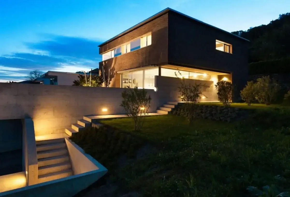 Moderna casa de dos plantas al anochecer, con iluminación exterior cálida, escaleras de piedra y un césped.