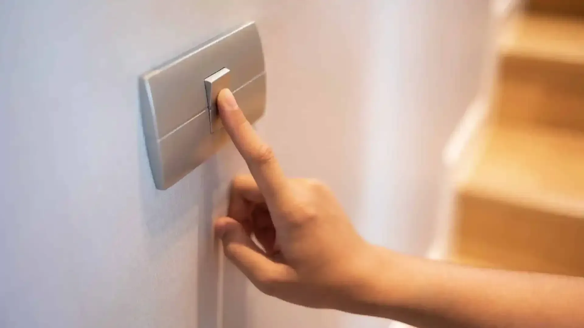 Primer plano de una mano pulsando un interruptor de luz rectangular plateado montado en una pared blanca.