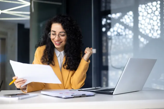 Un profesional sonriente, sentado en un escritorio con un ordenador portátil y documentos, celebrando con el puño en alto.