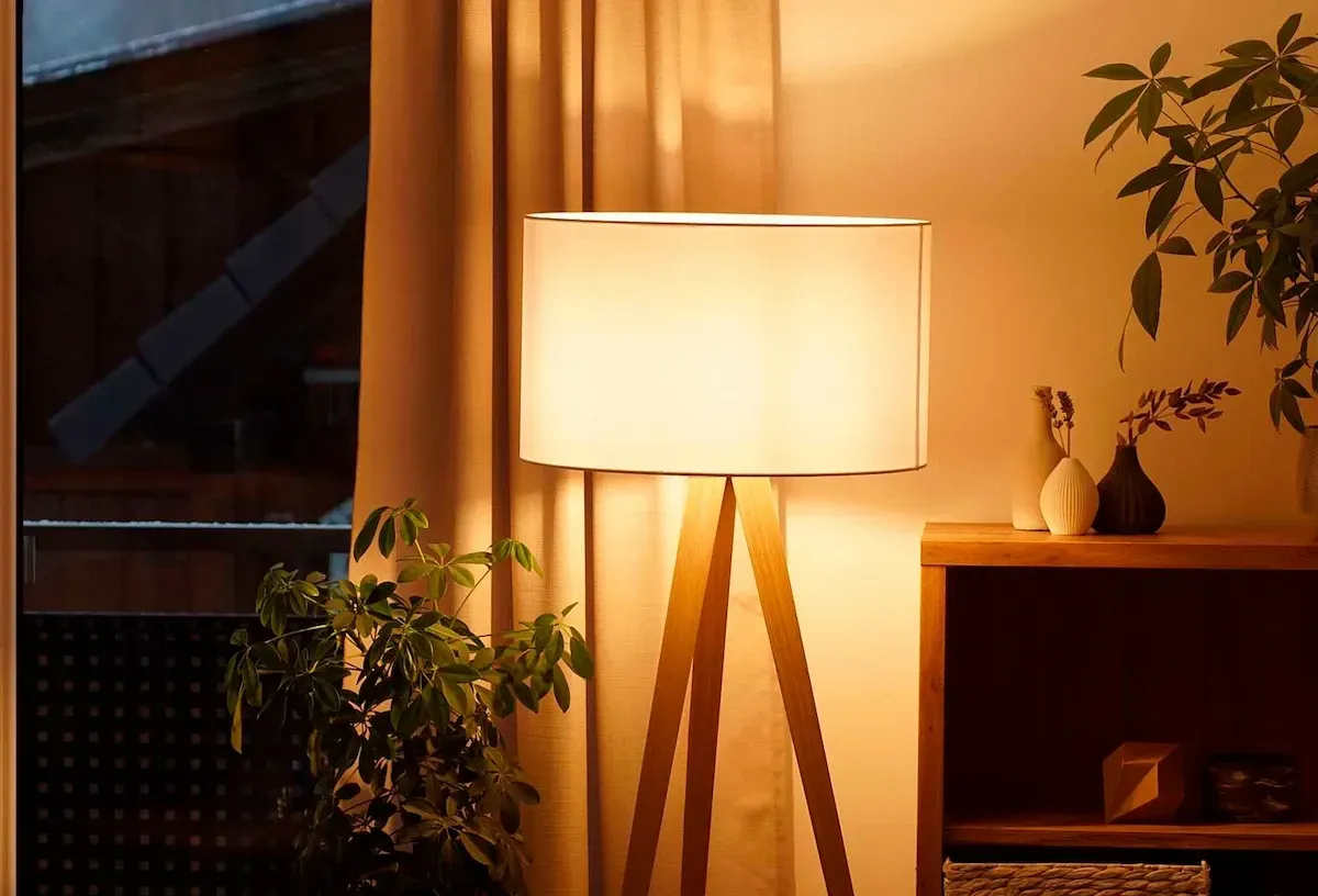 Una lámpara de pie con trípode y pantalla beige emite una luz cálida en una habitación con plantas de interio.