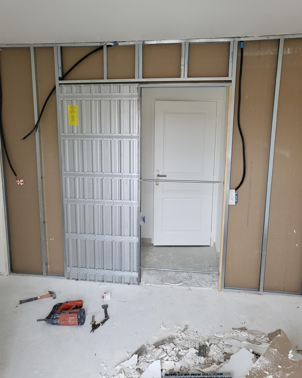 Chantier de construction avec ossature métallique pour une porte coulissante. On aperçoit des câbles électriques, des plaques de plâtre et une ossature partiellement construite.