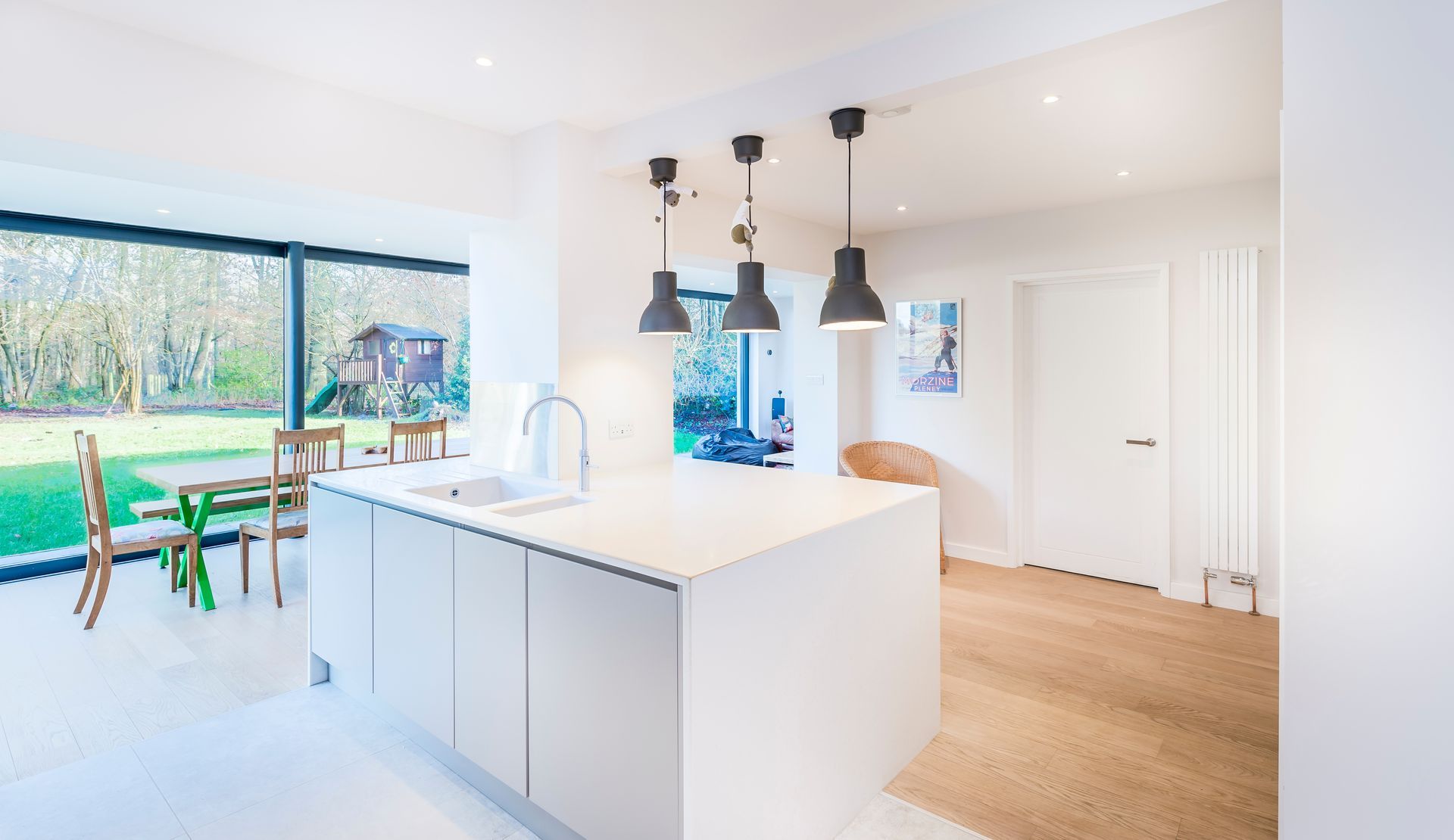 Cuisine blanche moderne avec îlot central, suspensions lumineuses et grande fenêtre donnant sur une cour.