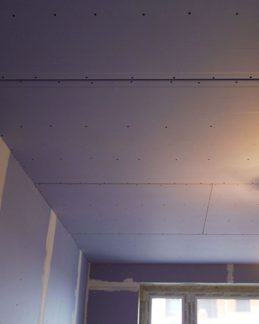 Plafond en plaques de plâtre avec trous de vis apparents, murs violet clair et fenêtre dans une pièce.