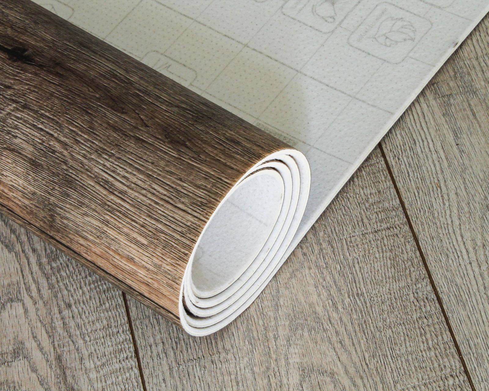 Rouleau de revêtement de sol vinyle imitation bois déroulé sur un sol similaire. On aperçoit le verso du rouleau avec les instructions.