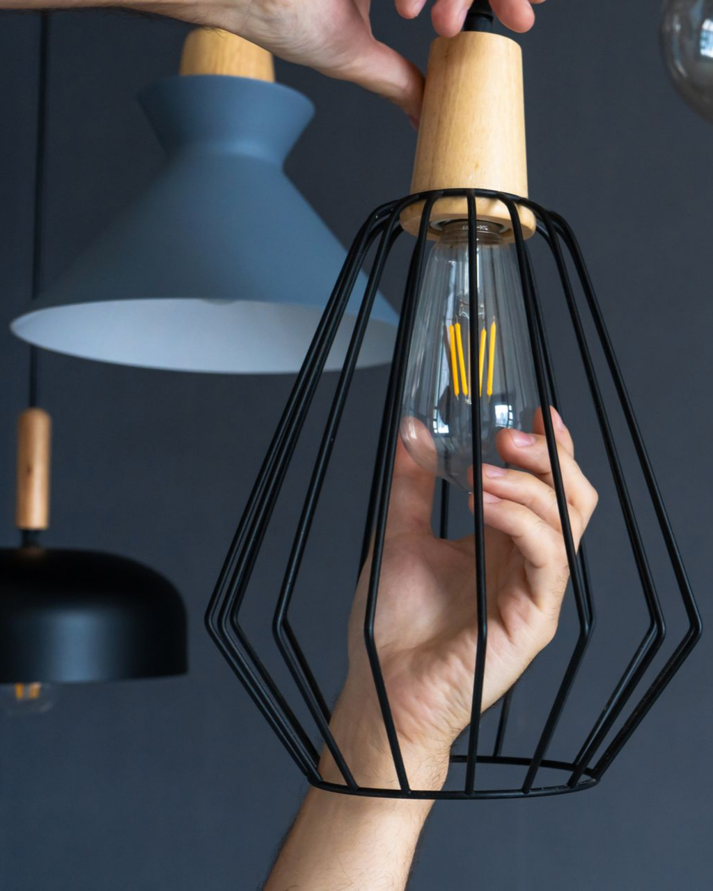 Une personne installe une ampoule dans une suspension lumineuse à structure métallique noire ; d'autres suspensions sont visibles en arrière-plan.