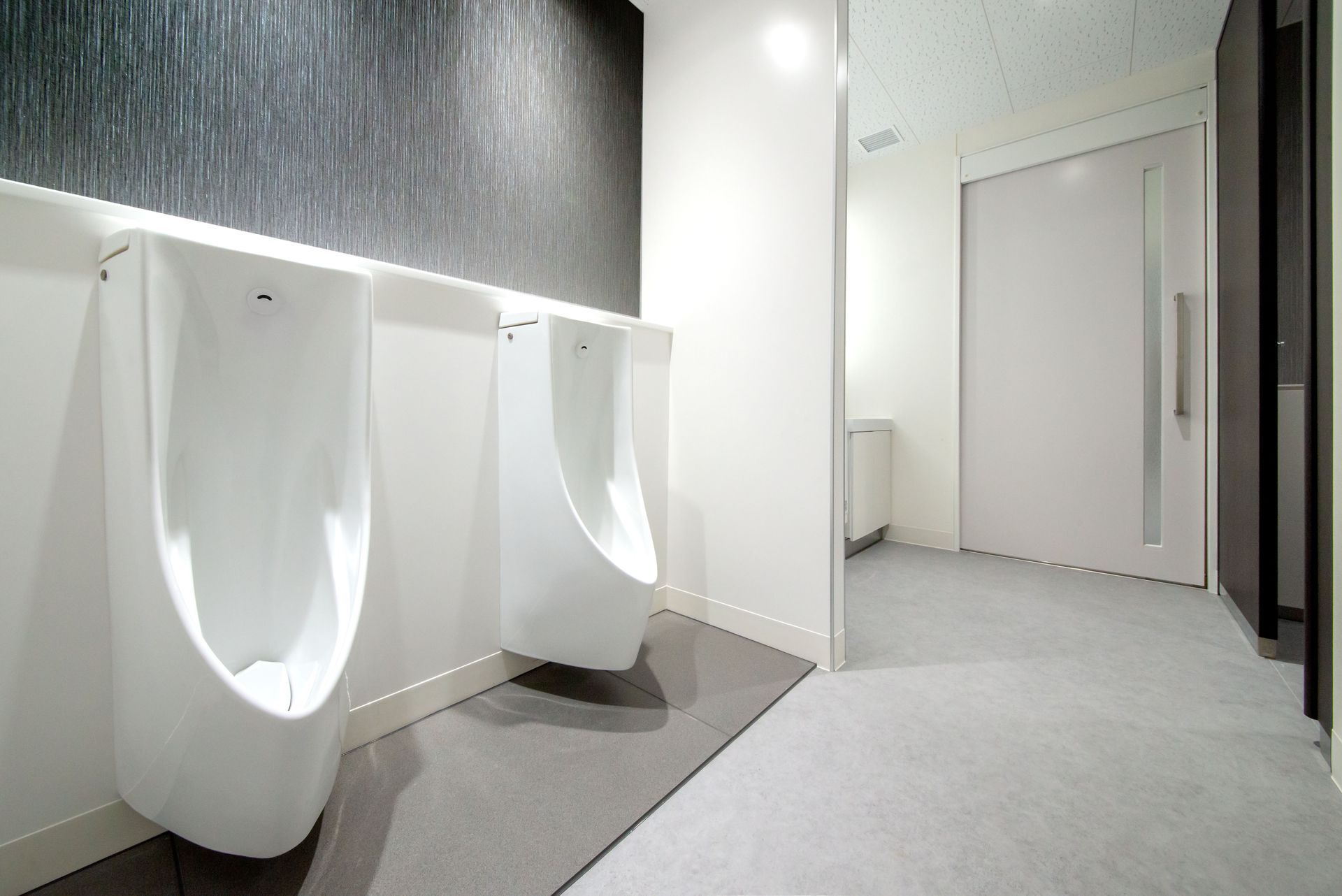 Deux urinoirs blancs dans des toilettes modernes pour hommes, avec un tapis gris et une porte.