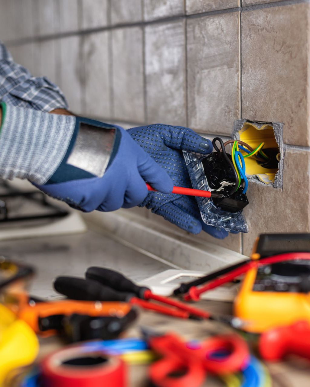 Électricien travaillant sur une prise murale, à l'aide d'un tournevis. Gants bleus, outils visibles, fond carrelé.