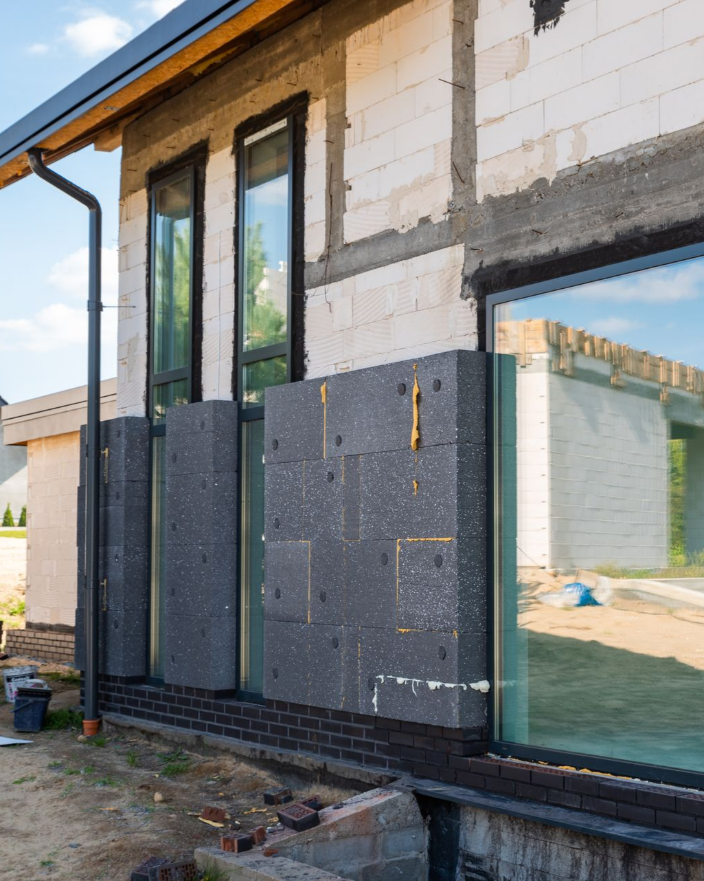 Vue extérieure d'un bâtiment en construction, montrant les panneaux d'isolation entre les fenêtres et les fondations en briques.
