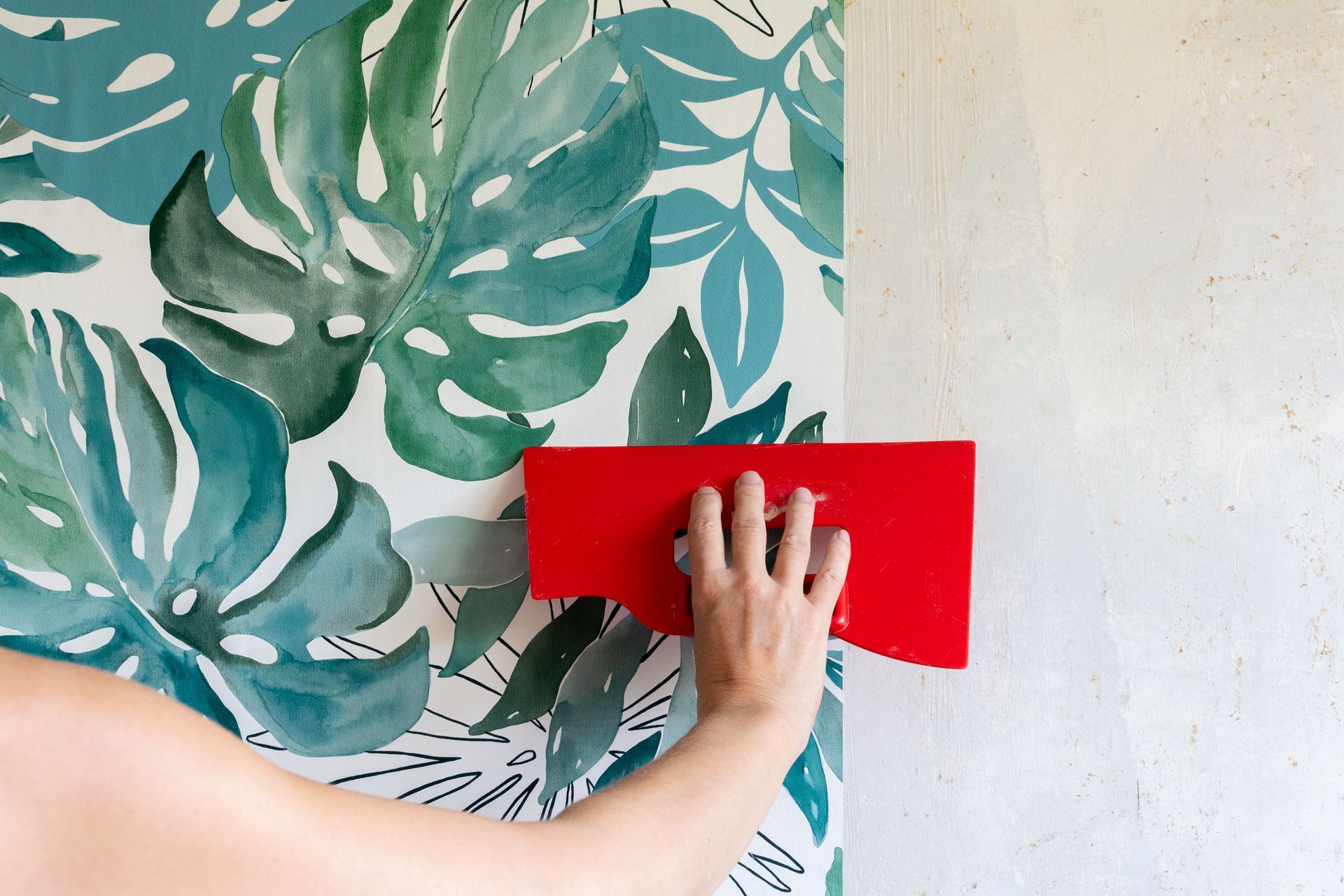 Une personne utilise un outil de lissage rouge pour appliquer un papier peint à motifs de feuilles aquarelles sur un mur blanc.