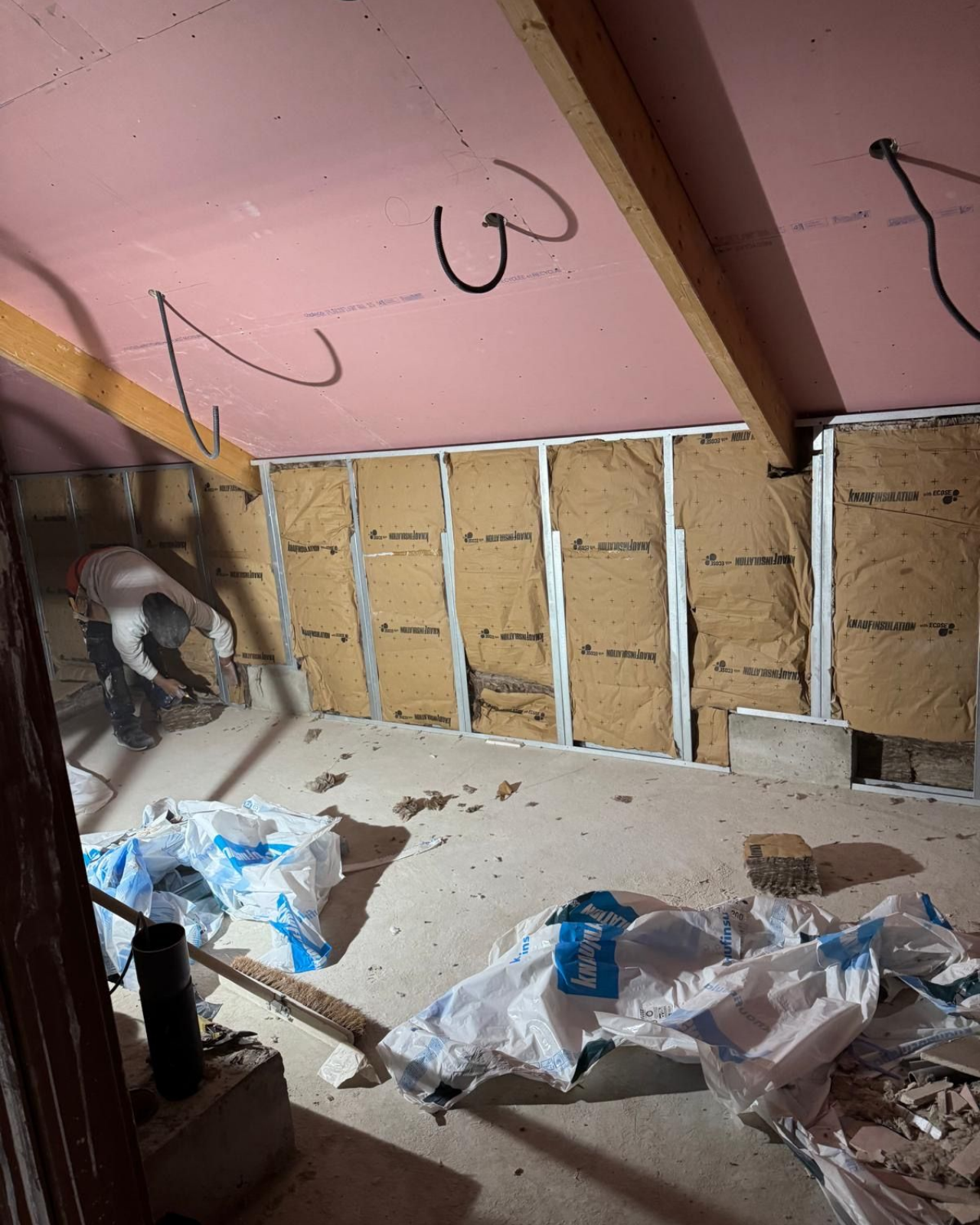 Travaux intérieurs : une personne installe de l’isolant entre les montants muraux ; plafond rose. Sacs de gravats au sol.