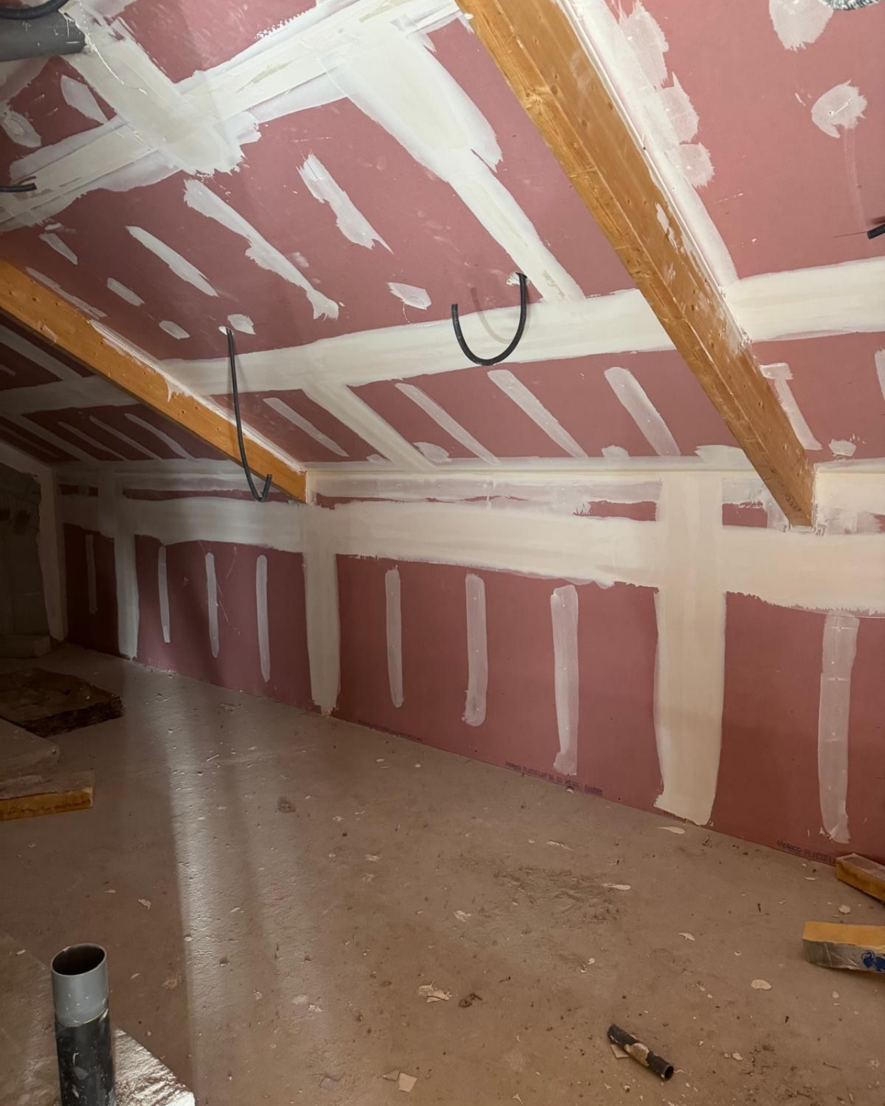 Intérieur du grenier avec cloisons sèches roses et enduit blanc. Poutres en bois et câblage électrique apparents.