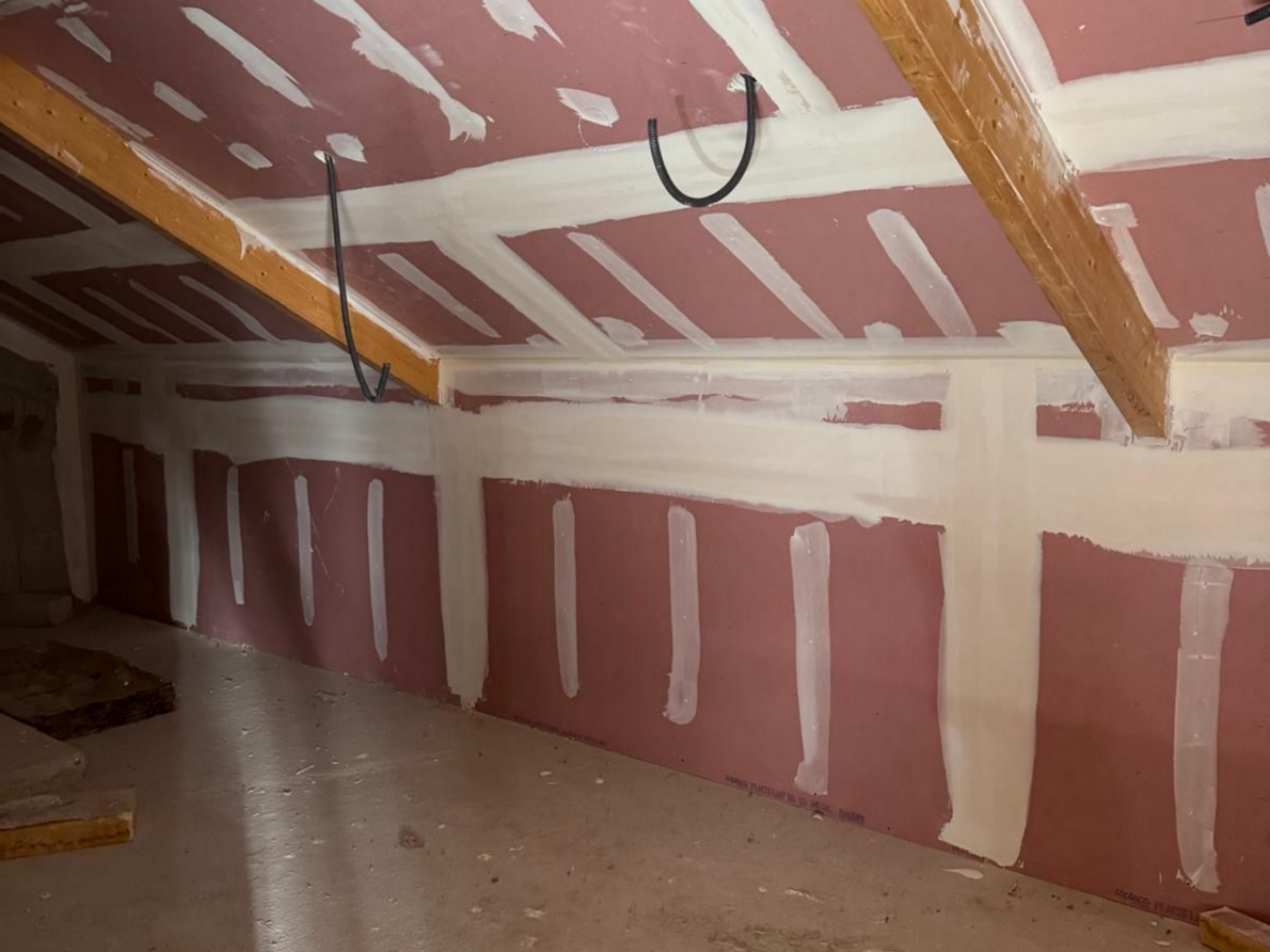 Vue intérieure d'un grenier avec des plaques de plâtre roses et des poutres en bois. Un enduit à joints blanc a été appliqué.