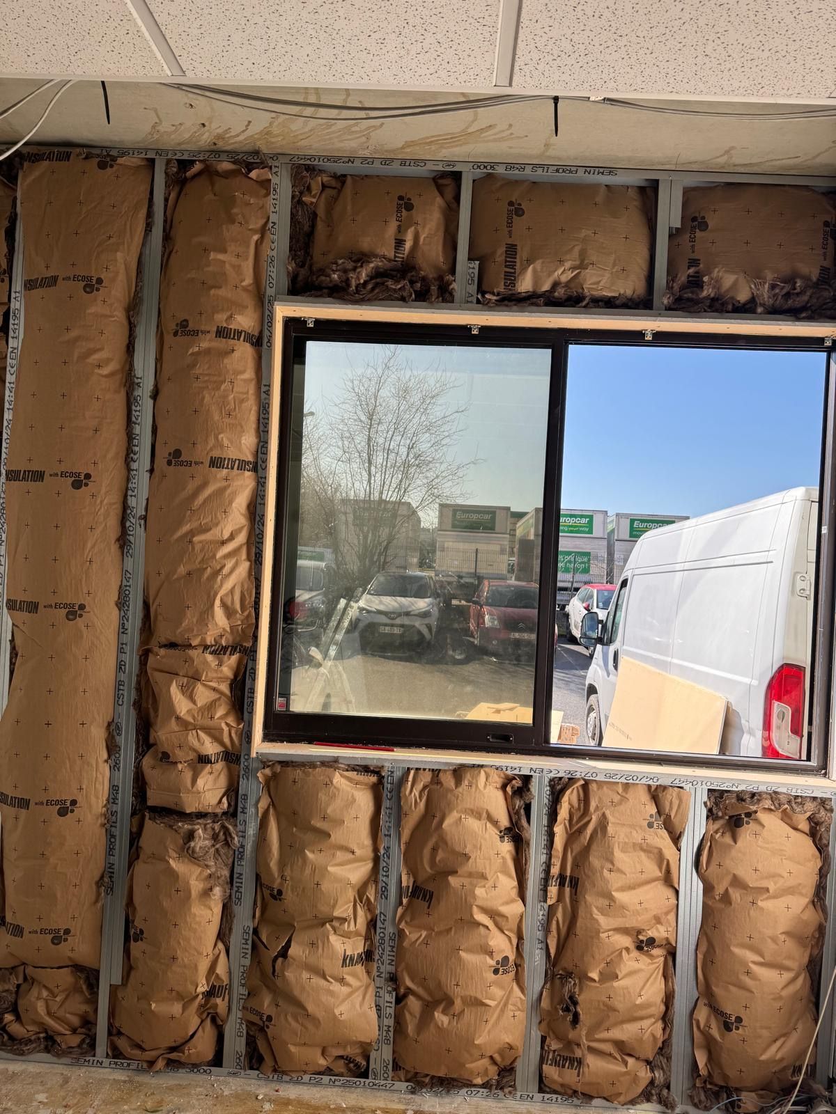 Mur en construction avec fenêtre ; isolation visible. Vue extérieure : véhicules et ciel.