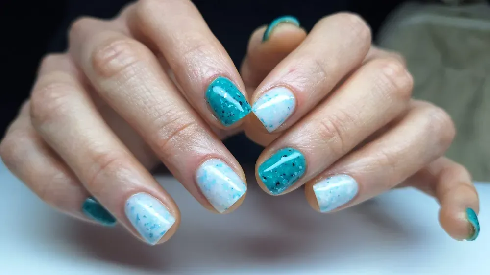 Manos con esmalte de uñas moteado en verde azulado y blanco.