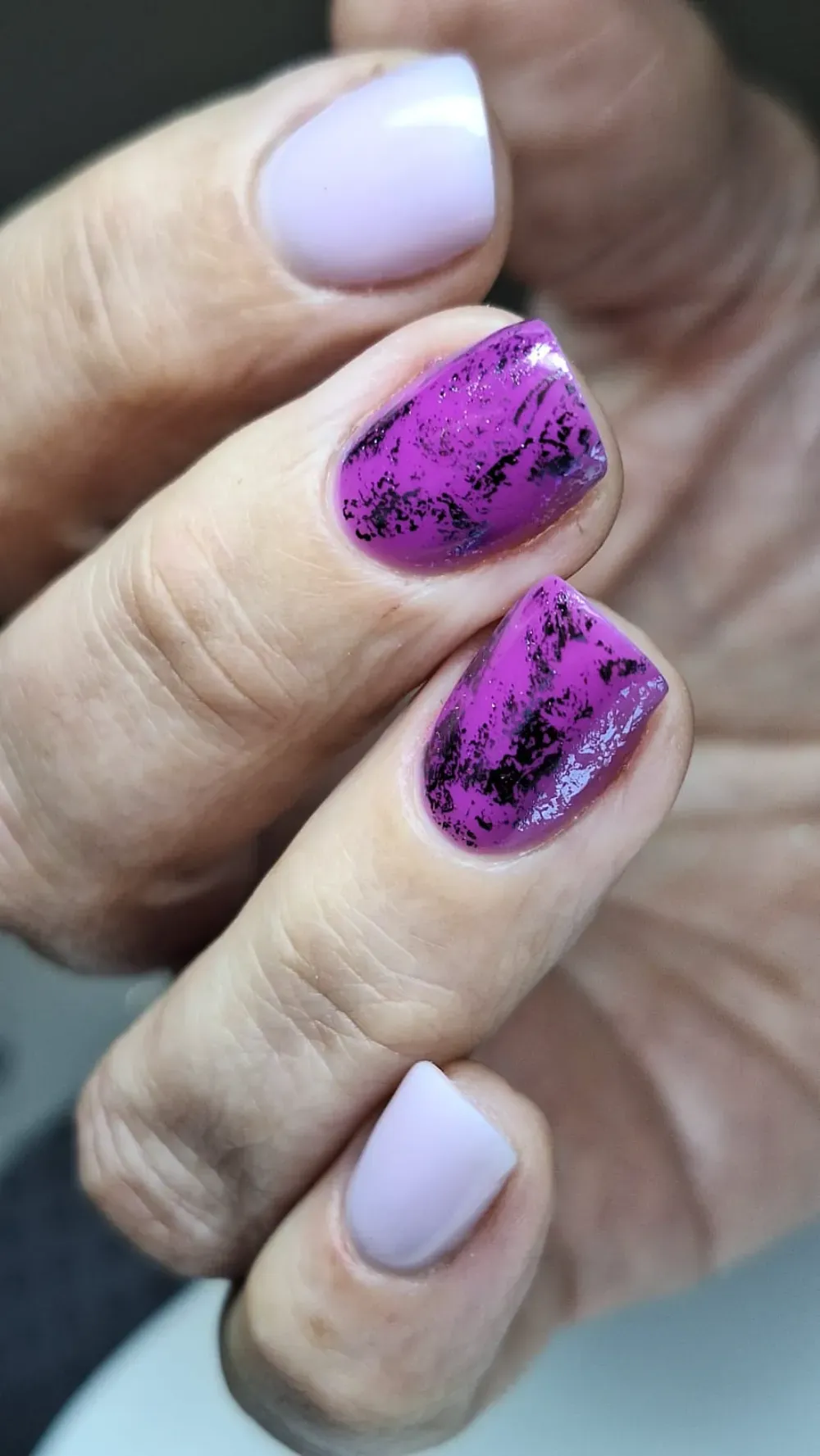 Mano con dos uñas de color violeta claro y dos uñas de color magenta con diseño de salpicaduras negras.