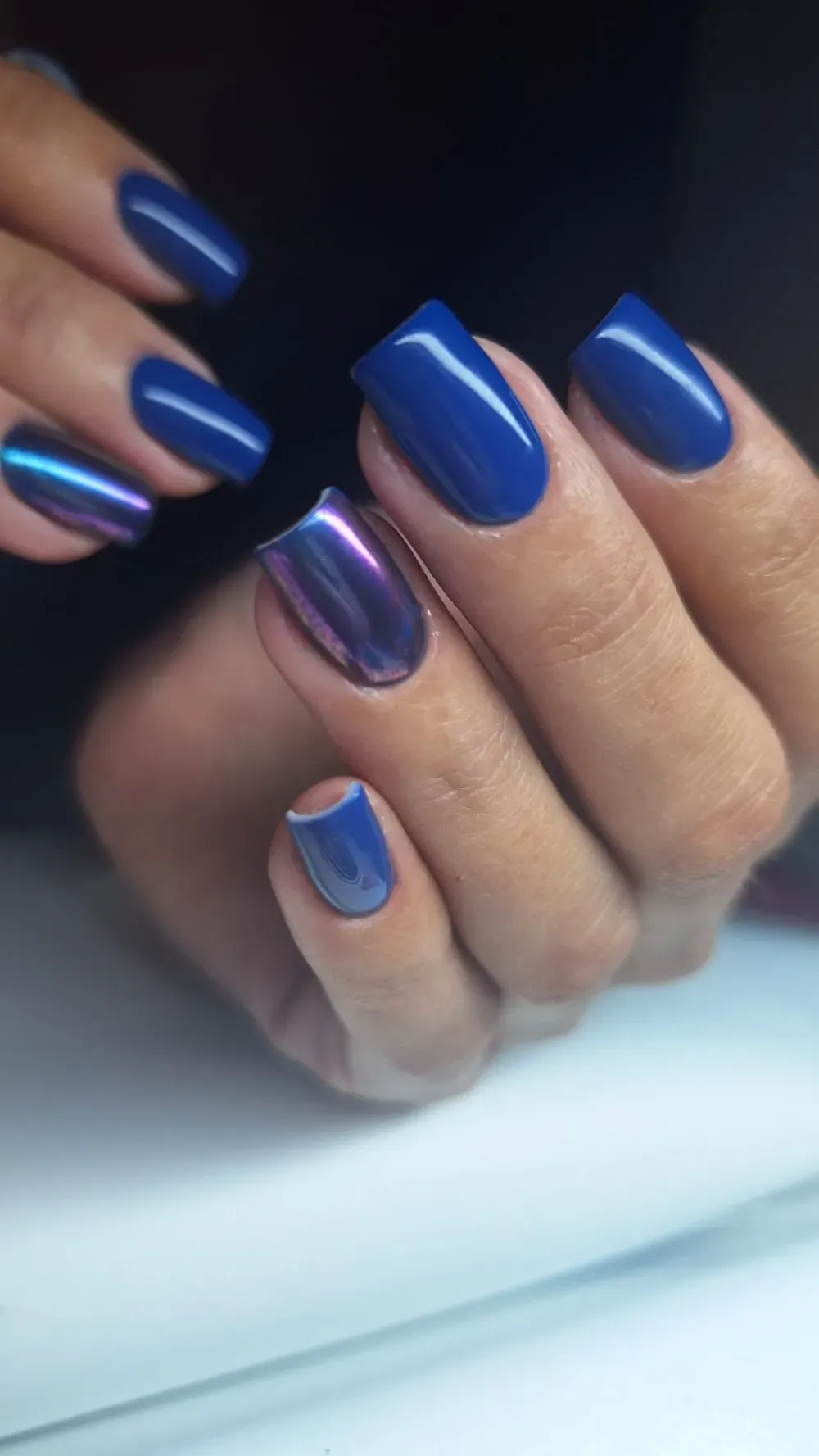 Manos con manicura azul, una uña acentuada con efecto cromado iridiscente.