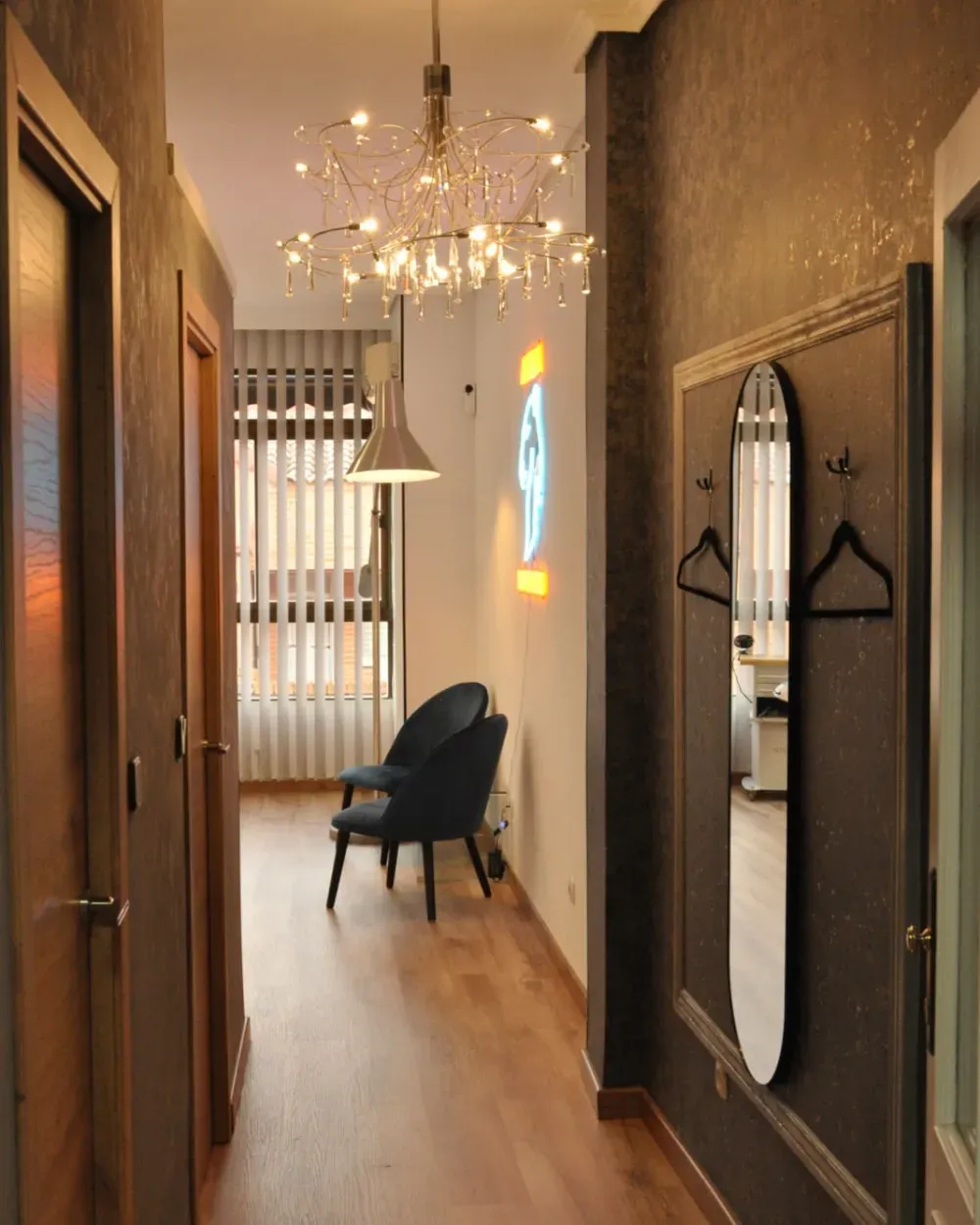 Pasillo con pisos de madera, paredes oscuras texturizadas, lámpara ornamentada, sillas y espejo.