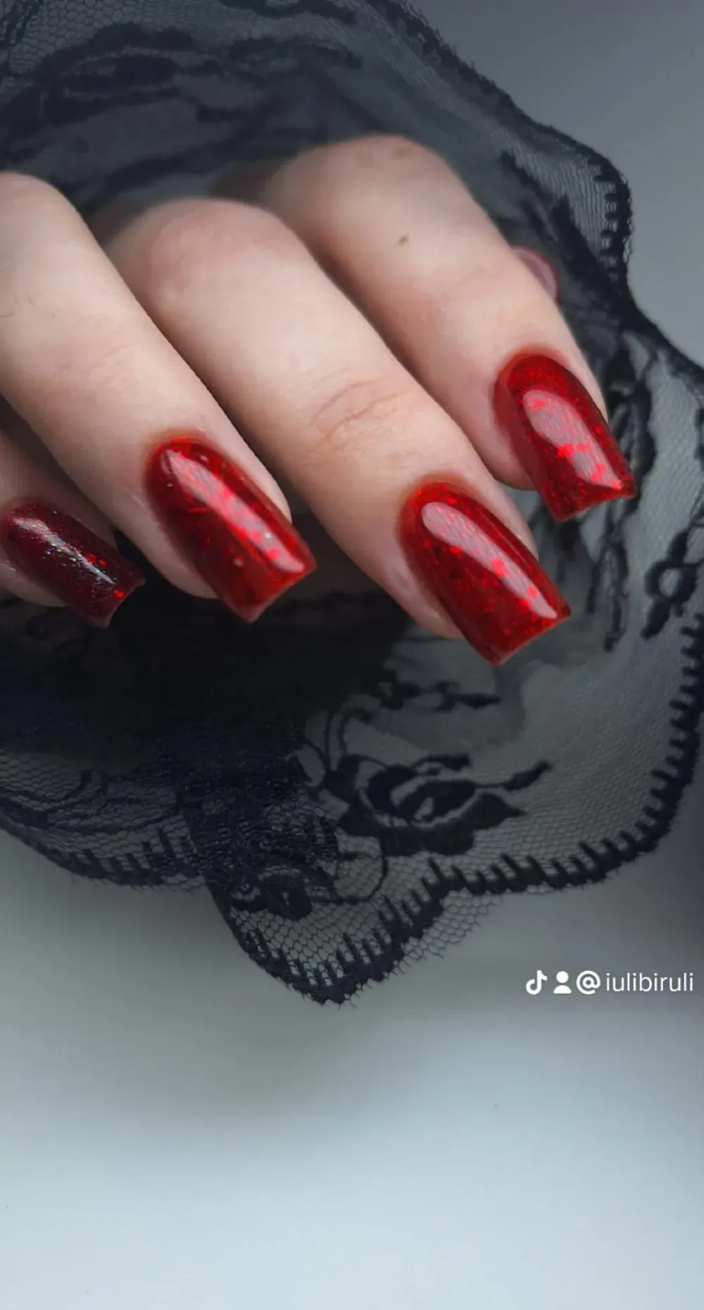 Mano con uñas rojas y cuadradas, apoyada sobre encaje negro, mostrando el brillo del esmalte de uñas.