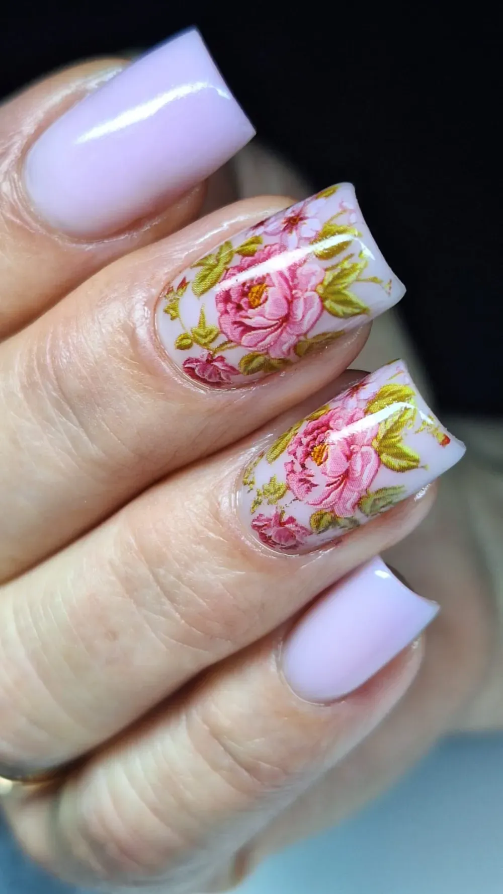 Uñas cuadradas de color morado claro, dos de ellas con calcomanías florales rosas.