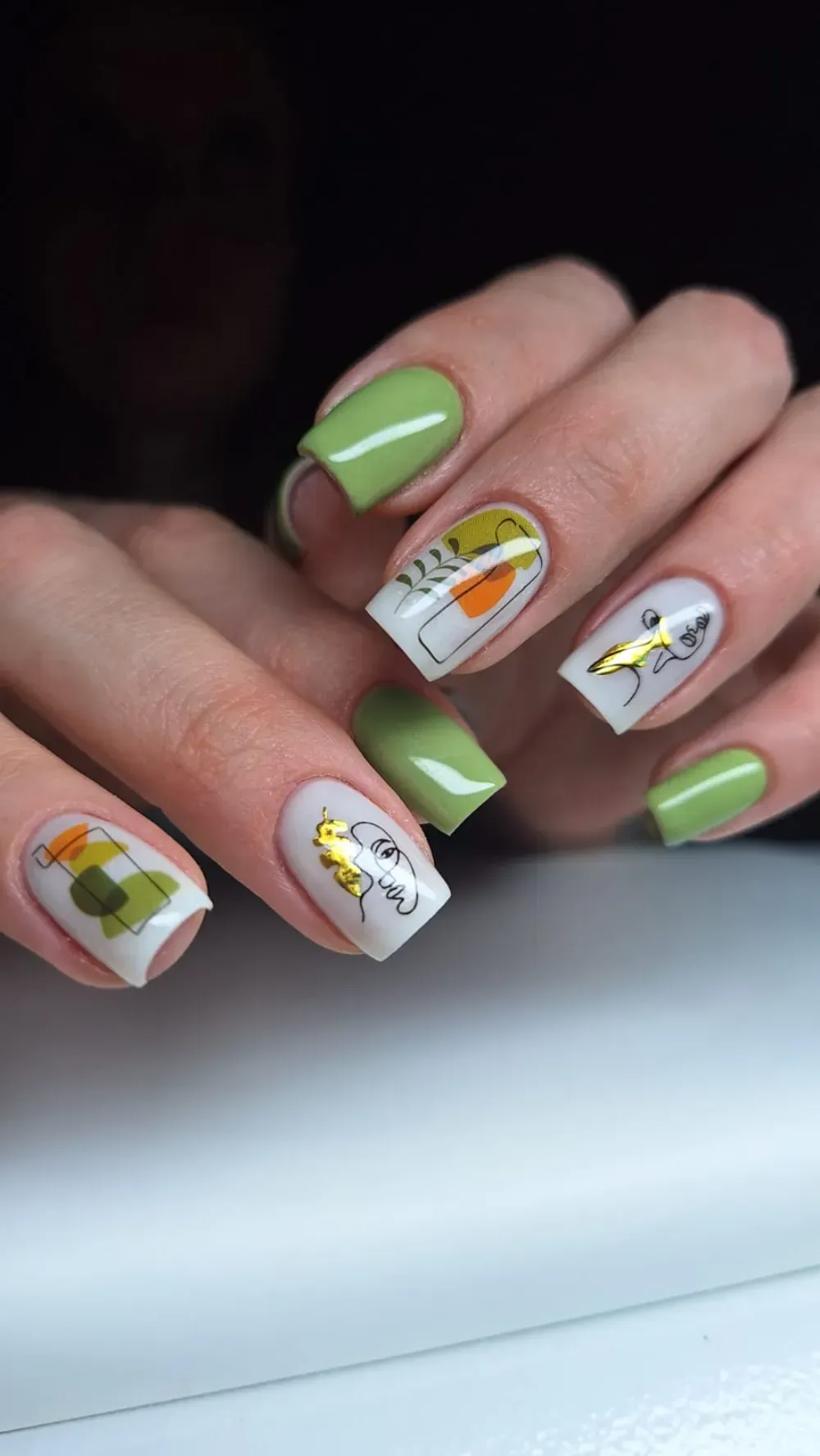 Manos con uñas cuadradas; esmalte verde en algunas, blanco con diseños abstractos de hojas de oro en otras.
