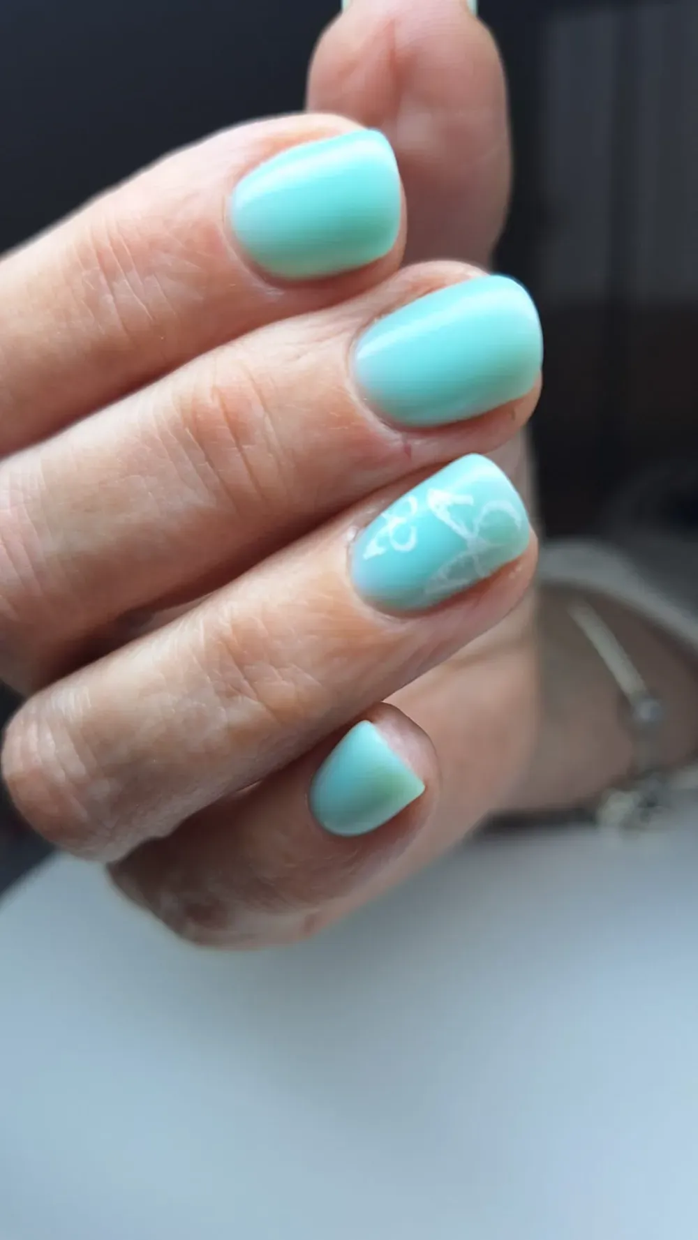 Mano con uñas cortas y redondeadas pintadas de turquesa claro. Una de ellas luce un diseño floral blanco.