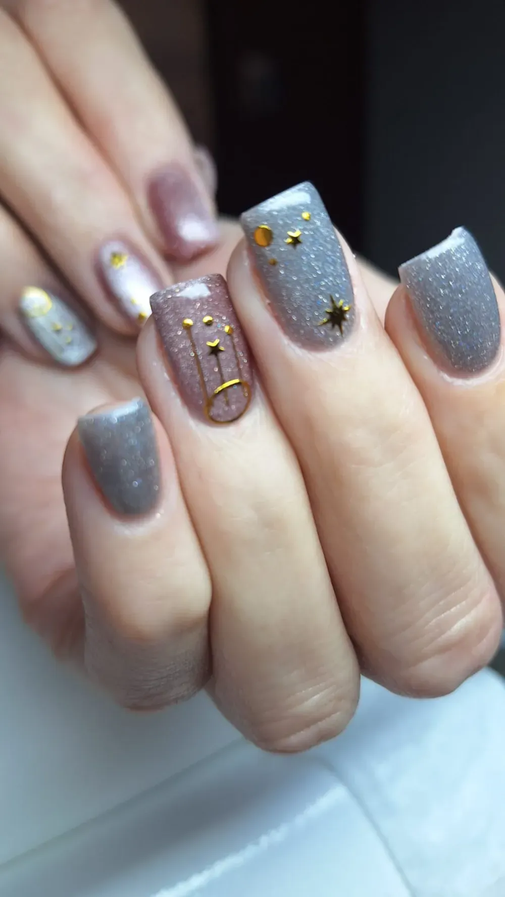 Primer plano de una mano con uñas cuidadas: tonos plateados y rosados ​​brillantes con acentos de estrellas y líneas doradas.
