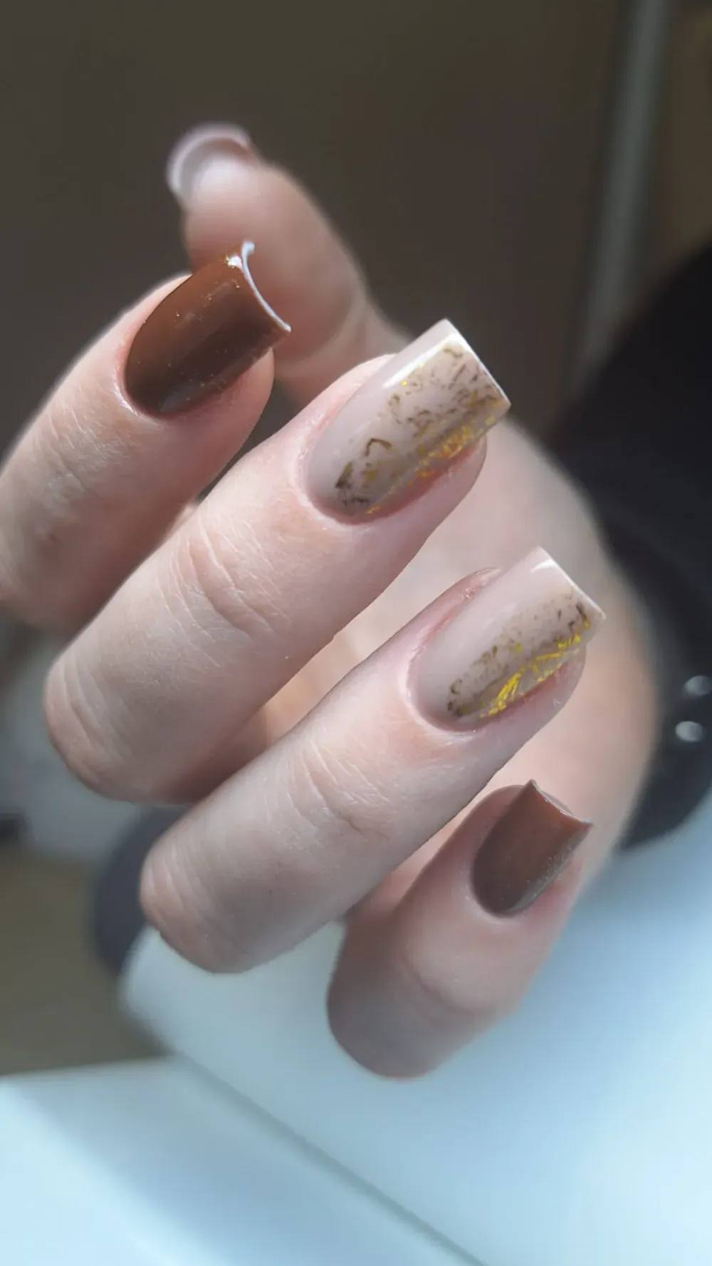 Mano con uñas de forma cuadrada; dos de color marrón, dos de color beige con vetas doradas y una con astillas blancas.