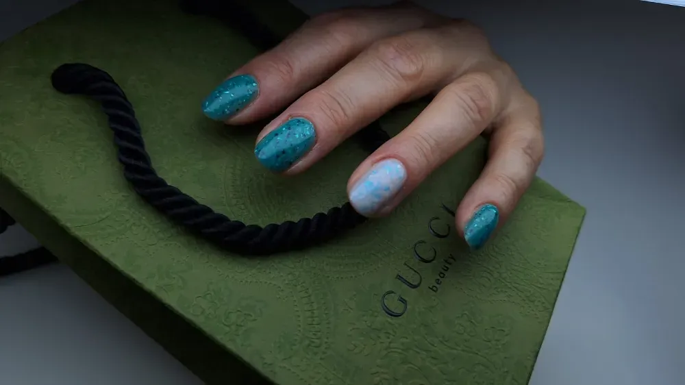 Una mano con uñas con brillantina color verde azulado reposa sobre una bolsa de compras verde de Gucci.