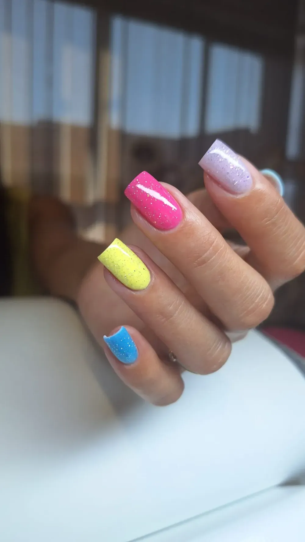 Mano con uñas cuadradas pintadas de azul, amarillo, rosa y morado brillante.