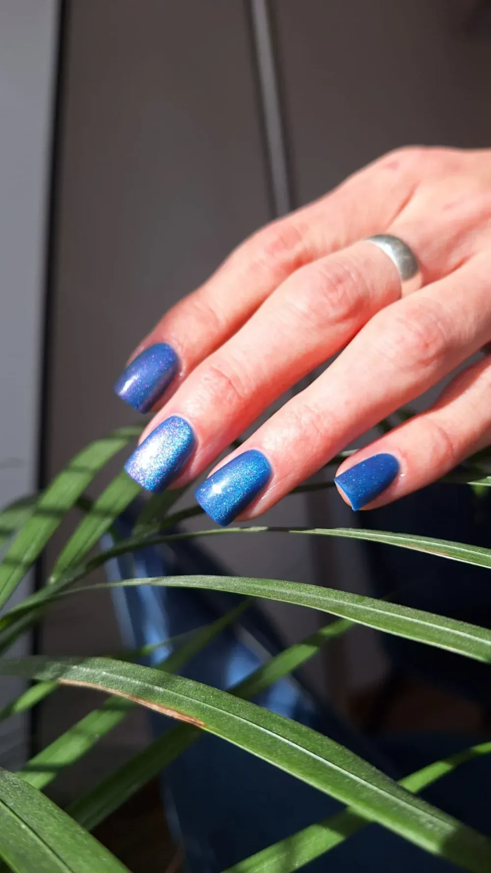 Mano con esmalte de uñas azul brillante, apoyada sobre hojas de plantas verdes.