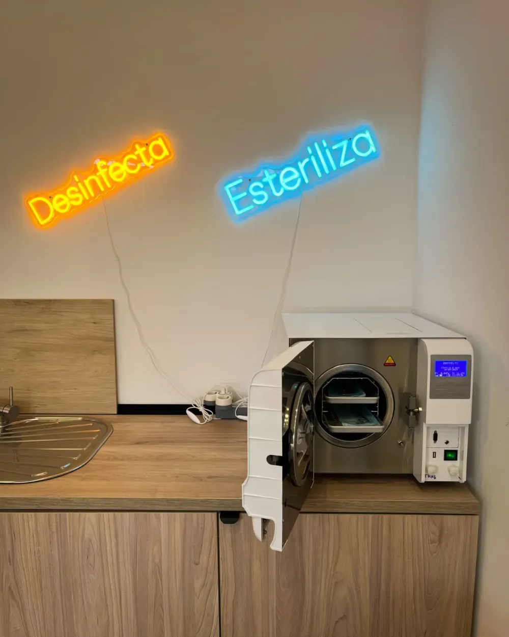 Autoclave con la puerta abierta, debajo de los letreros neón “Desinfecta” (naranja) y “Esteriliza” (azul).