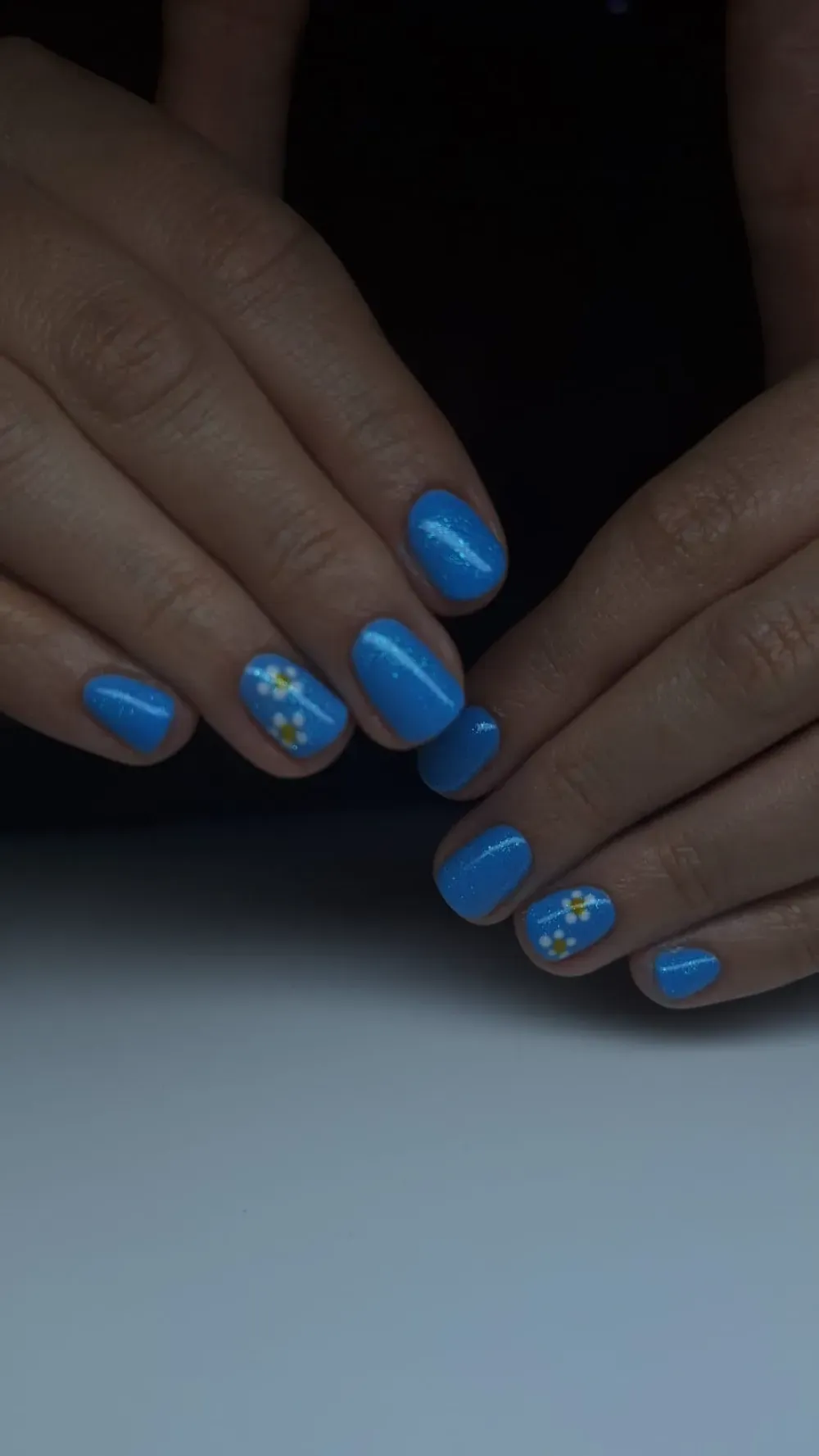 Manos con esmalte de uñas azul brillante, dos uñas con pequeños diseños de flores blancas.