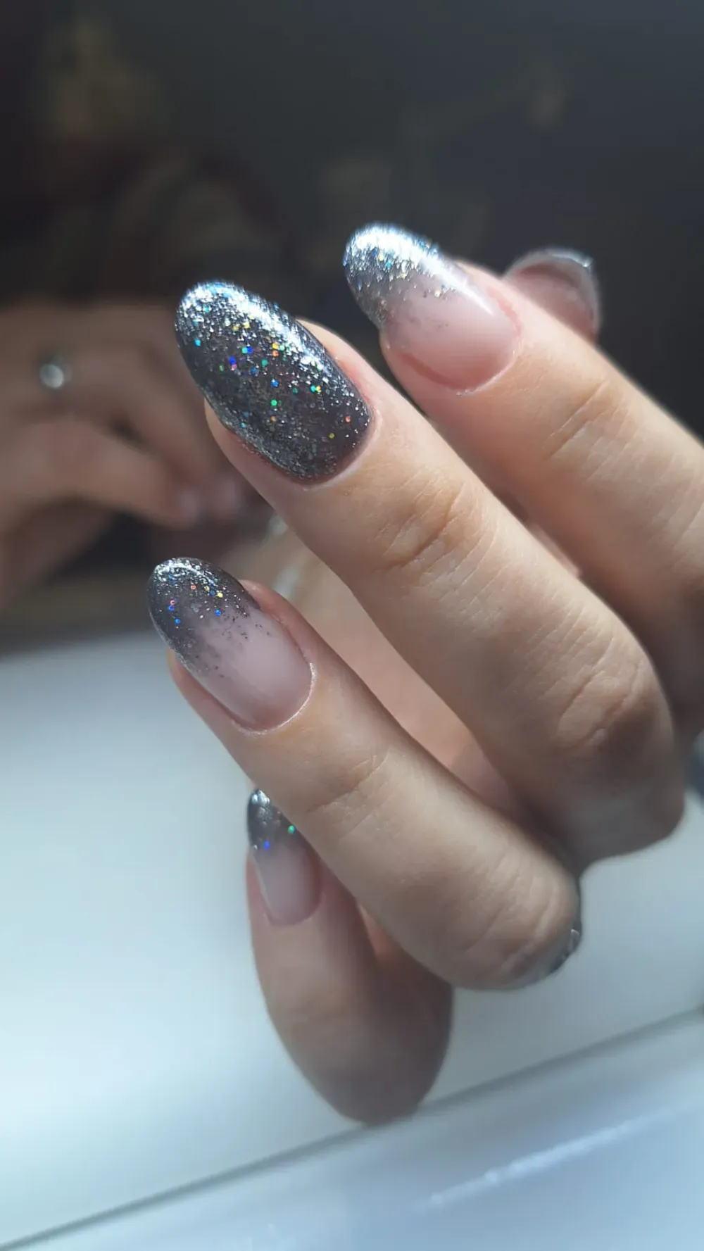 Mano con uñas de forma ovalada que presenta un diseño ombré con brillo plateado.