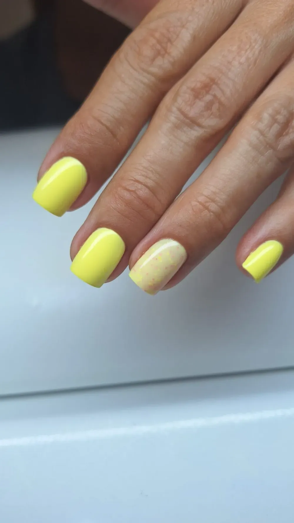 Mano con esmalte de uñas amarillo, una uña con un tono más claro, sobre una superficie blanca.
