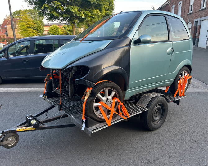 Une voiture bleu clair à deux portes, endommagée à l'avant, se trouve sur une remorque, fixée avec des sangles orange.
