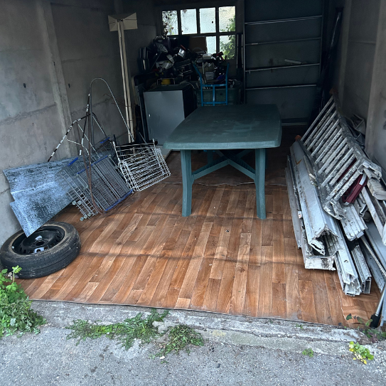 Abri de rangement encombré avec une table verte, des objets en métal et un pneu sur un sol à motifs de bois.