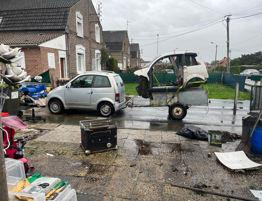 Petite voiture argentée tirant une remorque avec un châssis de voiture dans une rue encombrée et humide.