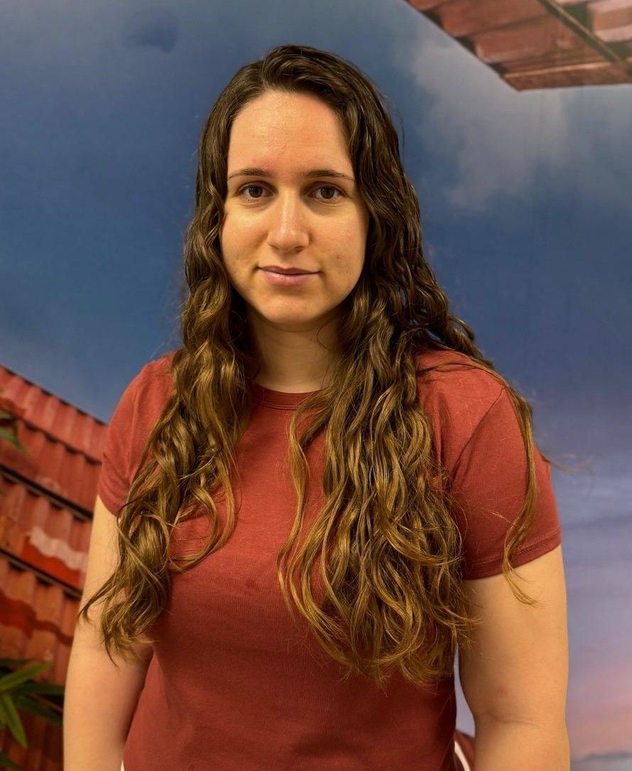 Un hombre con camisa roja sonríe, apoyado en una mesa y con reloj. Al fondo se ve un cielo azul y parte de un edificio.