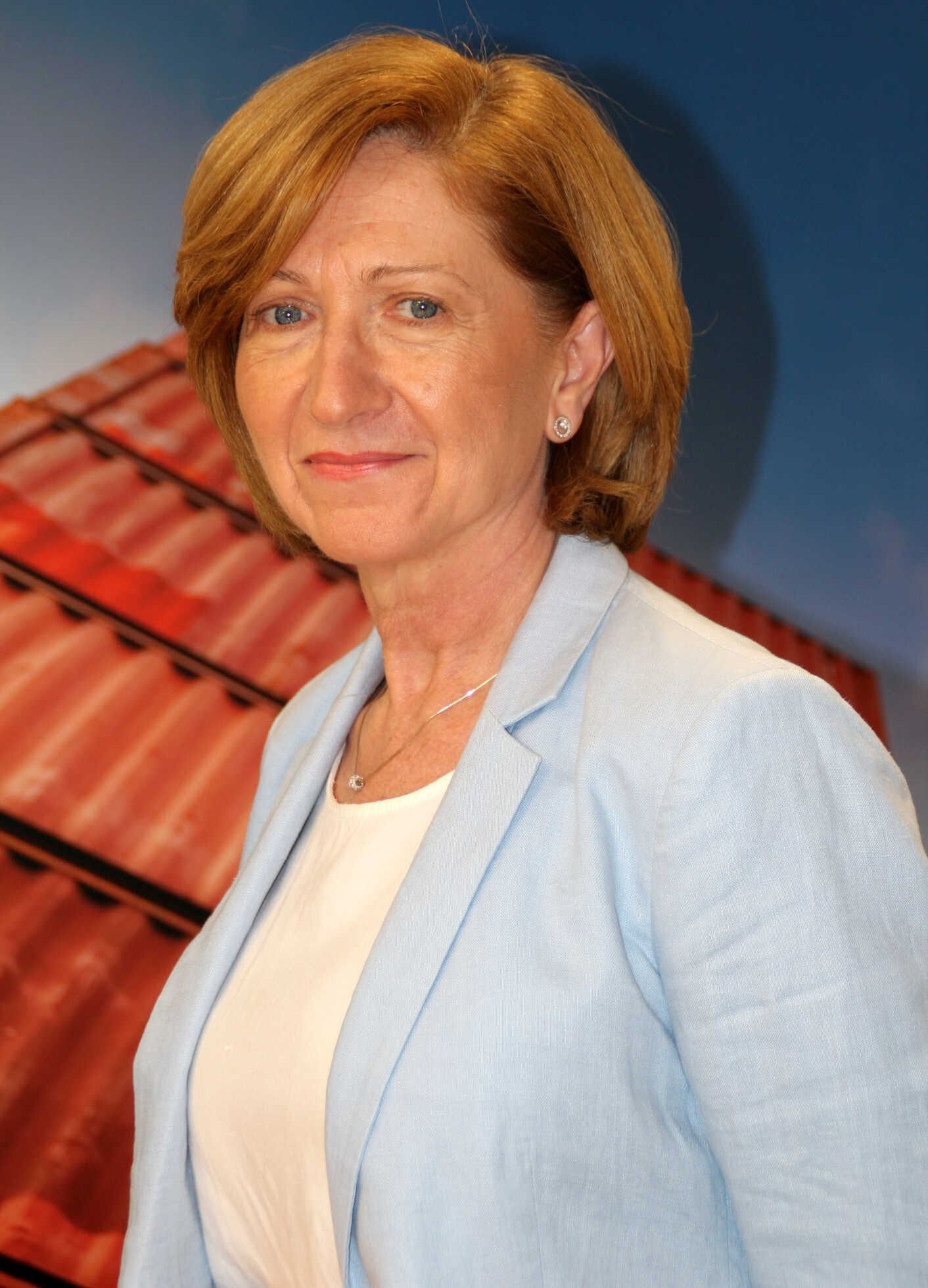 Mujer con una chaqueta azul claro, sonriendo, frente a un tejado de tejas de fondo.