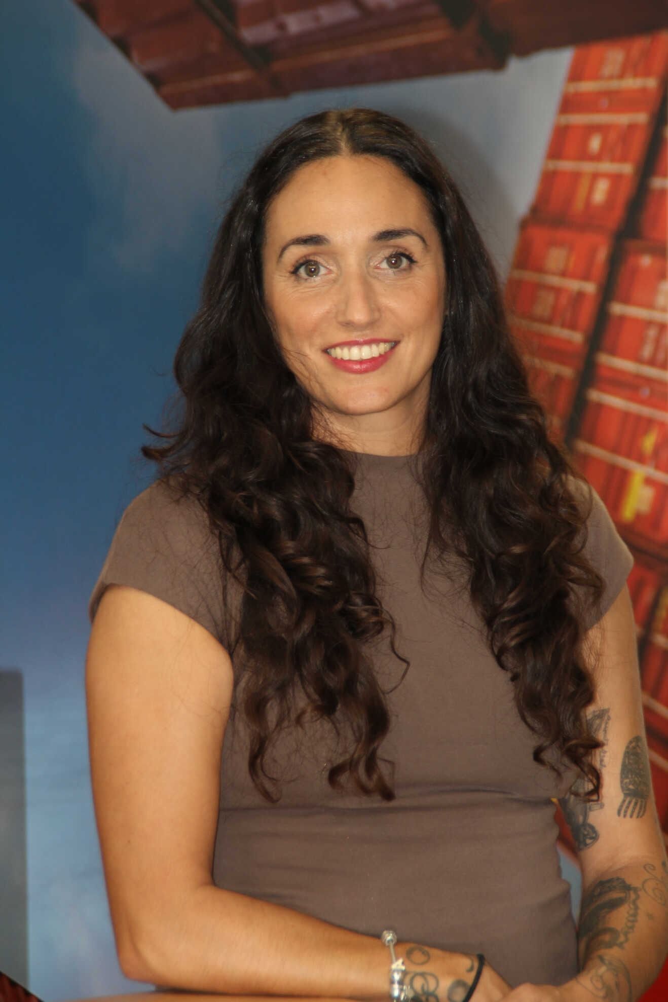 Mujer con cabello largo y oscuro, parte superior marrón, sonriente, tatuaje en el brazo, delante de un fondo colorido.