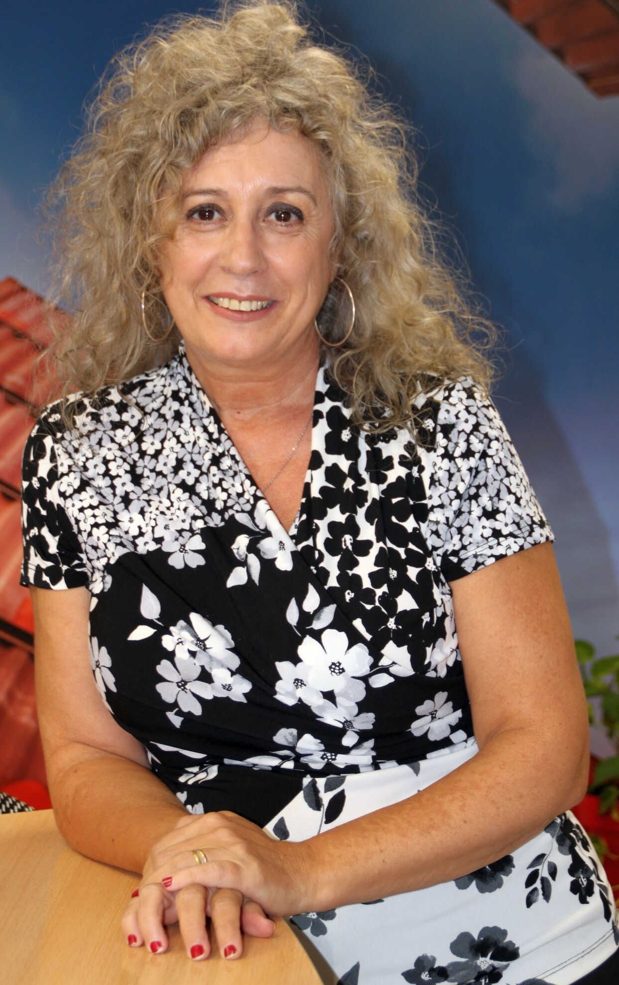 Mujer con cabello gris rizado con un top floral blanco y negro, sonriendo y apoyando los brazos sobre un objeto marrón.