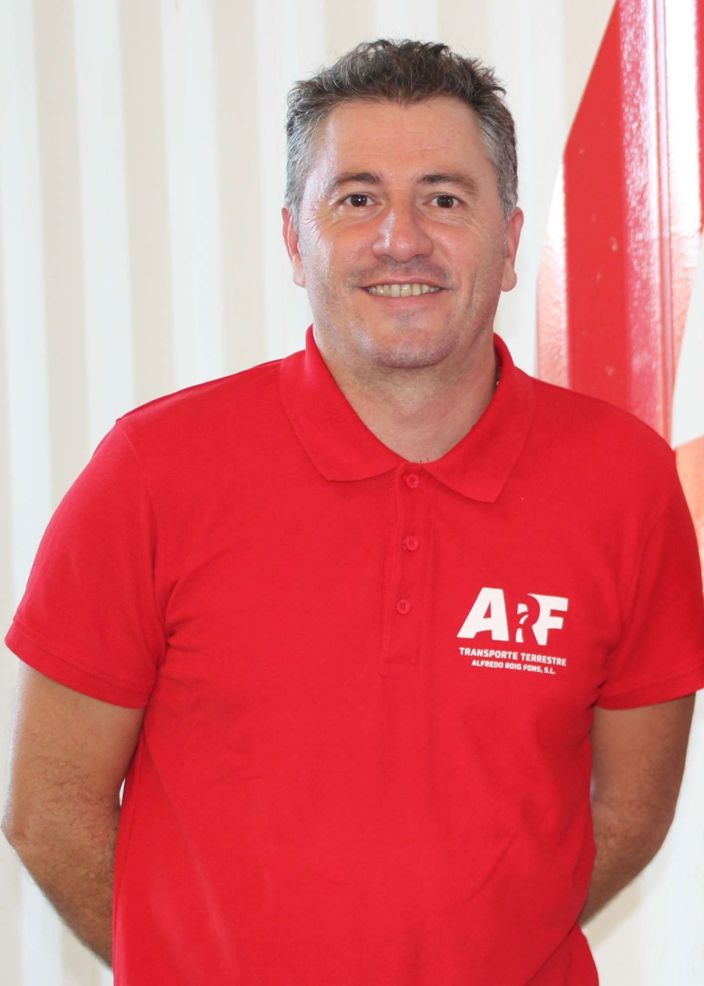 Un hombre con una camisa polo roja sonríe a la cámara, de pie en el interior, cerca de un objeto rojo y blanco.