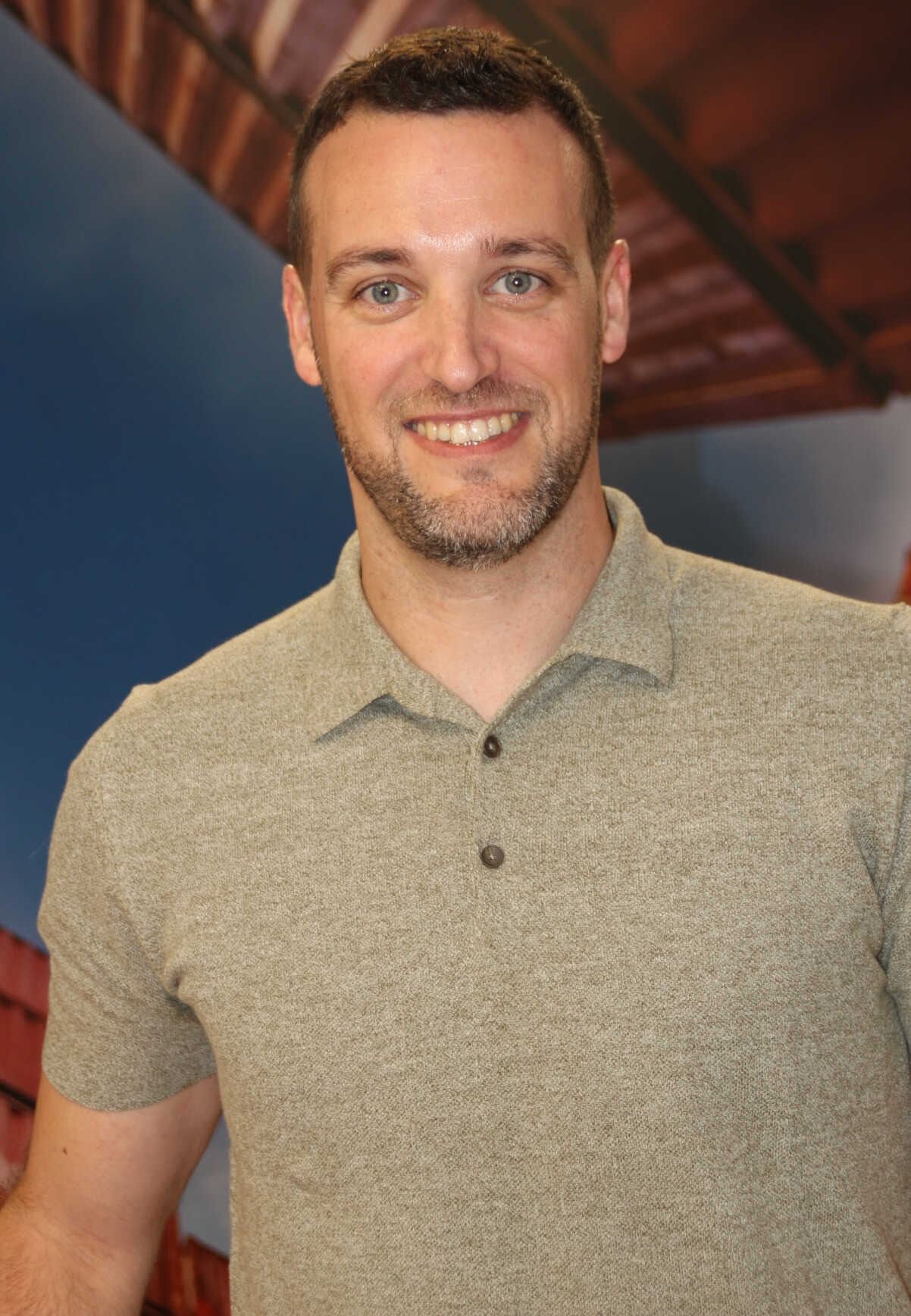 Un hombre con camisa roja sonríe, apoyado en una mesa y con reloj. Al fondo se ve un cielo azul y parte de un edificio.