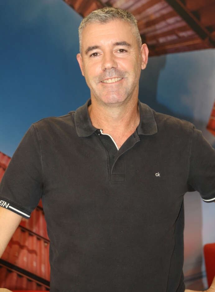 Un hombre con camisa roja sonríe, apoyado en una mesa y con reloj. Al fondo se ve un cielo azul y parte de un edificio.