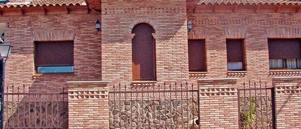 Una casa de ladrillo color canela con techo de tejas, entrada arqueada con dos columnas de piedra y palmeras a ambos lados.