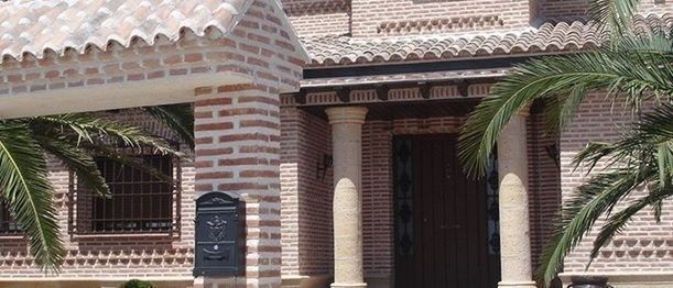 Una casa de ladrillo color canela con techo de tejas, entrada arqueada con dos columnas de piedra y palmeras a ambos lados.