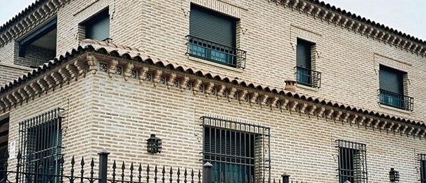 Un edificio de dos plantas construido con ladrillos de color claro, con varias ventanas con contraventanas oscuras y rejas metálicas.