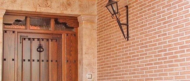 Una puerta de entrada de madera con listones verticales y una aldaba de metal oscuro enmarcada en piedra, junto a una pared de ladrillo con una lámpara.