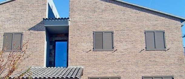 Un edificio de ladrillo de varias plantas con ventanas de contraventanas grises, una entrada central retranqueada y una sección de tejado de tejas.